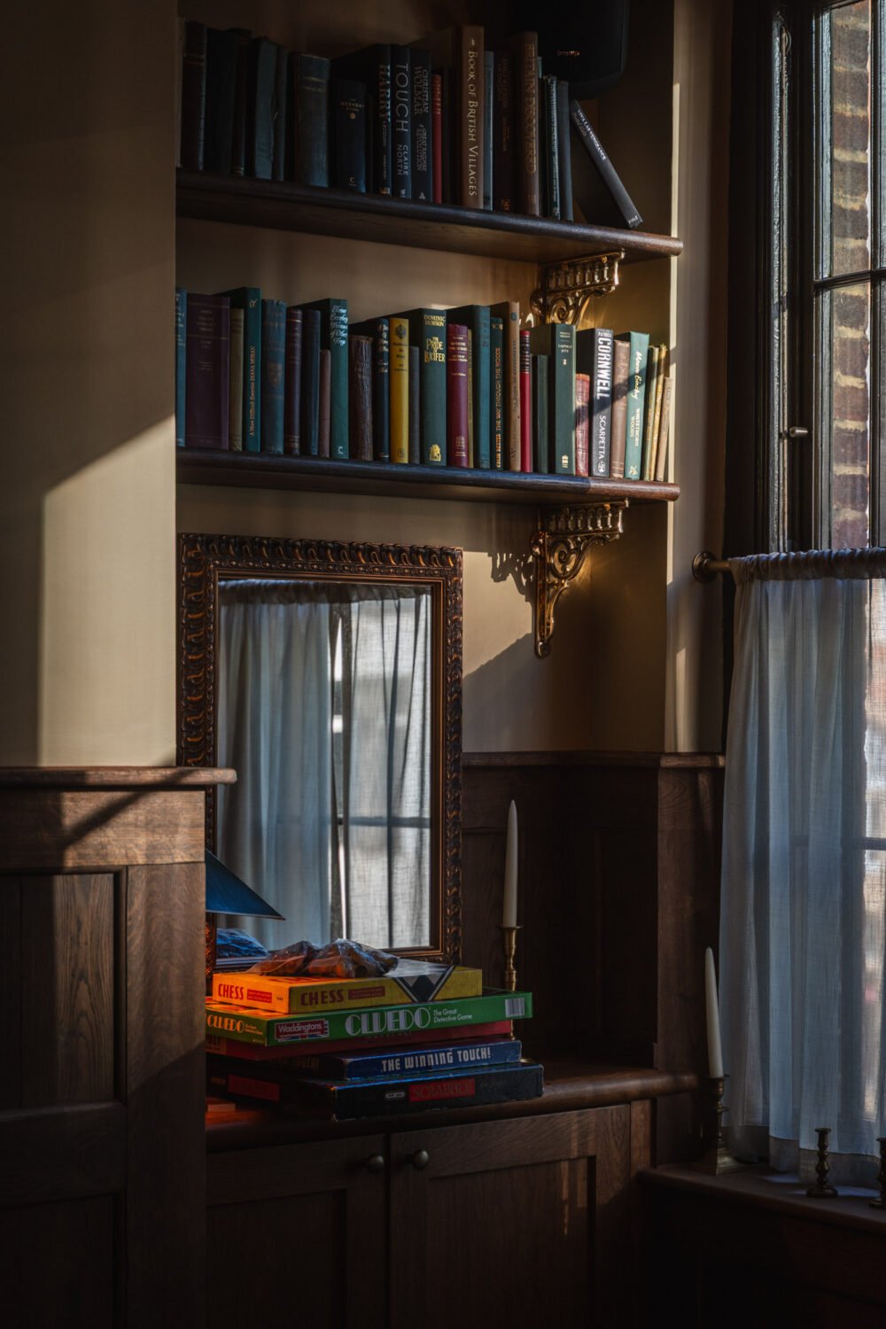 A character-filled pub corner with bookshelves, vintage mirror and warm natural light creating a cosy, lived-in atmosphere. Traditional London pub interior detail with wooden panelling, bookshelves, vintage mirror, board games and soft window light.