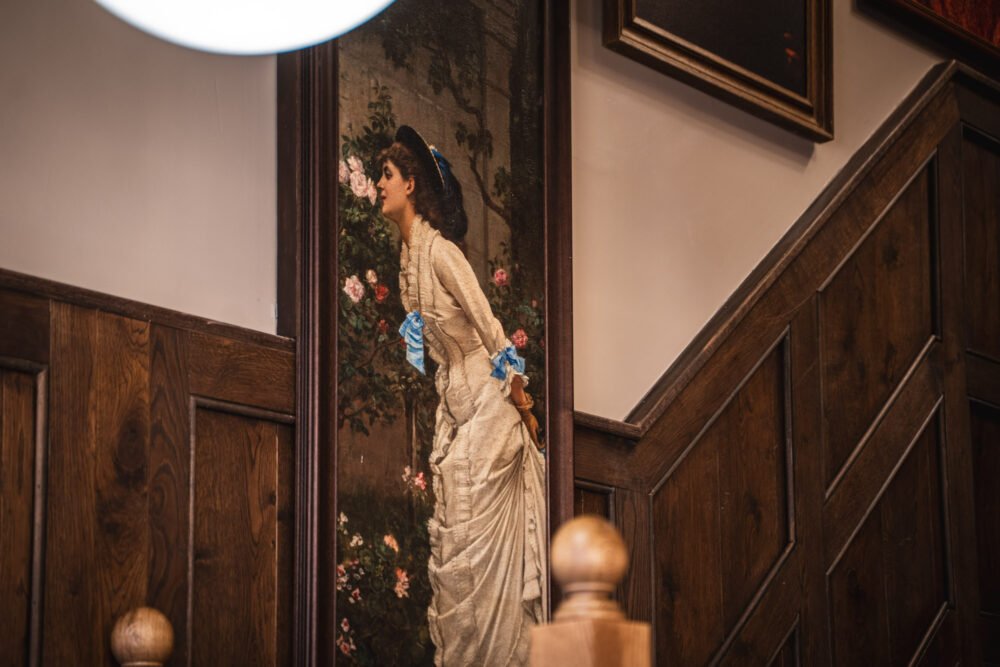 A characterful pub staircase with classic wood panelling and vintage artwork, adding depth and heritage to the space. Traditional London pub interior with wooden staircase and vintage framed artwork on panelled wall.