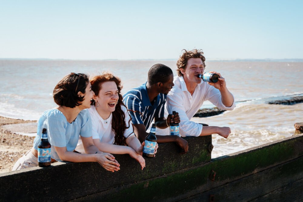 Friends enjoying Whitstable Bay 0.5% beer on a sunny day by the coast. Group of friends enjoying Whitstable Bay alcohol-free beer by the seaside