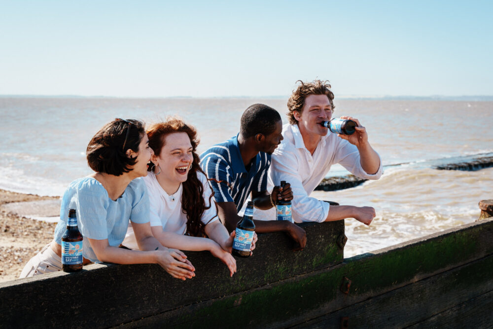 Friends enjoying Whitstable Bay 0.5% beer on a sunny day by the coast. Group of friends enjoying Whitstable Bay alcohol-free beer by the seaside